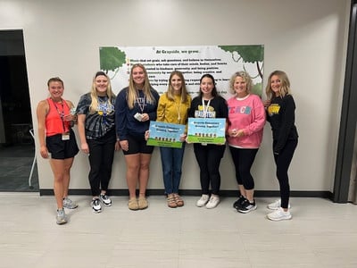 Seven teachers are standing against a wall holding two signs.