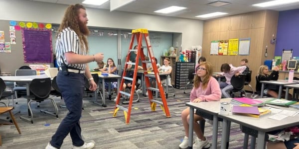 One adult talking to a classroom of students with a ladder in the middle of the room.