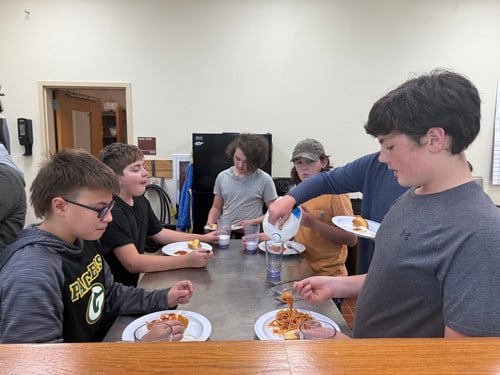 Six students eat spaghetti, with one student pouring milk in a glass.