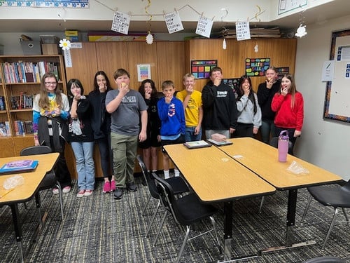 Students stand in a classroom taking a bite of an apple.