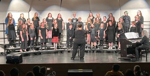 A middle school choir performs on stage, led by a conductor, for an audience.