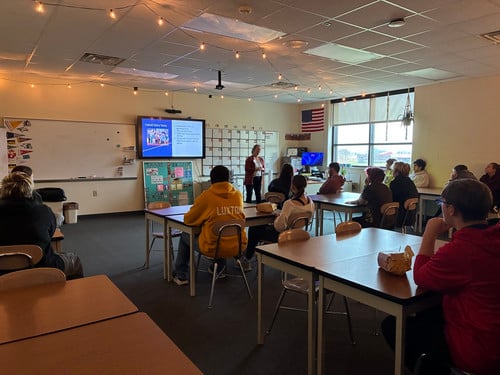 An adult presents a slide show to high school students seated at desks in a decorated classroom.