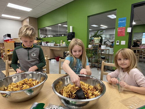 Three young students are mixing snacks in large silver bowls.