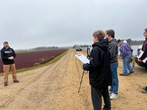 Students using a tripod and clipboard to interview a man at a cranberry bog.