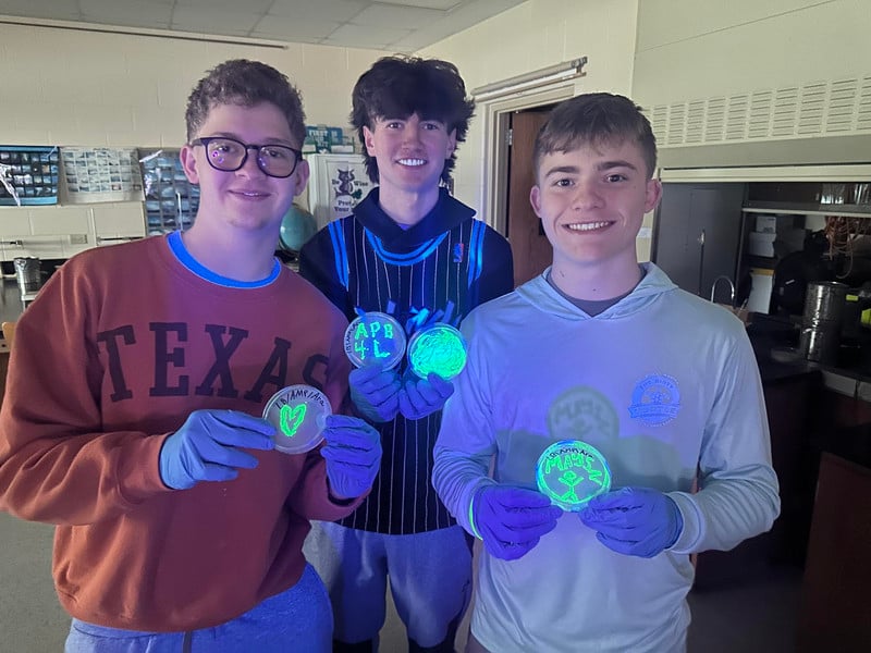 Three students in a science lab holding Petri dishes that glow bright neon green under UV light.