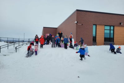 Preschoolers in colorful winter gear play on a large snowbank outside a brick school building.