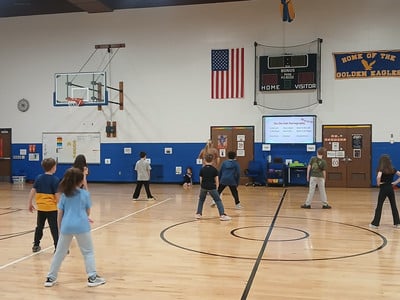 Students in a gym follow Cha Cha Slide choreography shown on a large screen during PE class.