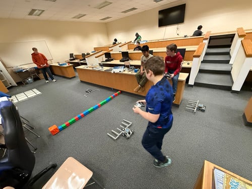 Students use remote controls to maneuver small robots around an obstacle course in a classroom.