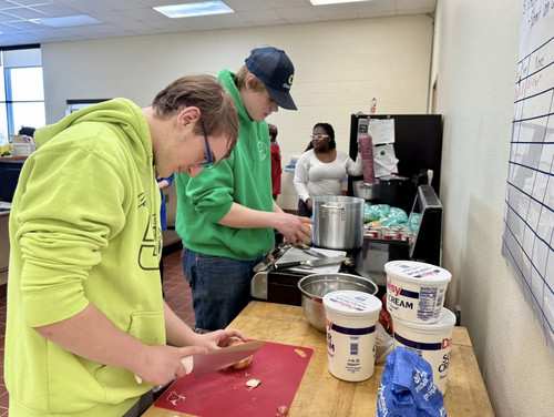 Two students in green hoodies chop onions and prepare food in a school kitchen.