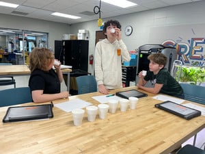 Three students conduct a milk taste test using several small white cups on a classroom table.
