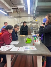 Students observe a demonstration of colorful liquid samples in test tubes on a metal lab table.