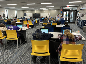 A classroom of students sits at individual desks working on laptops and wearing headphones.
