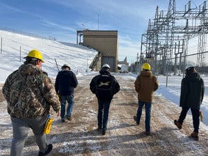 A group of people wearing hard hats walks toward a large industrial power facility in the snow.