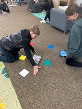 Two students sit in a hallway working together on a literacy project with papers and a laptop.