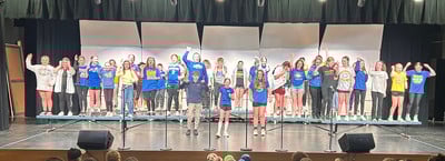 A large group of 6th-grade students stands on risers performing during a choir concert.