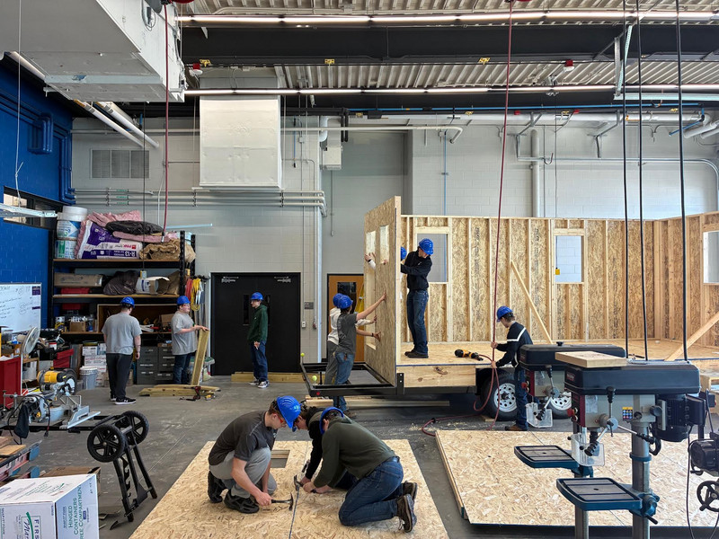 Students use power tools to construct the wooden frame of a tiny house in a workshop.