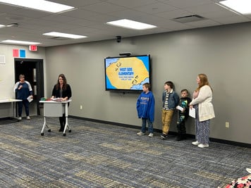 Ms. Routson presents at a podium to a school board with four people stand nearby in a meeting room.