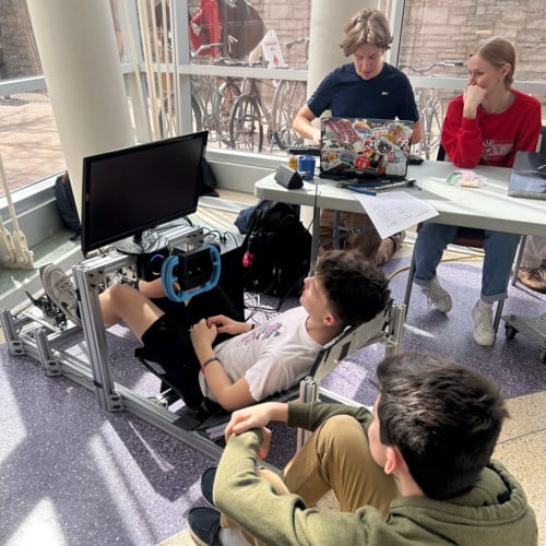 A student uses a metal racing simulator rig while other students watch in a sunny room.