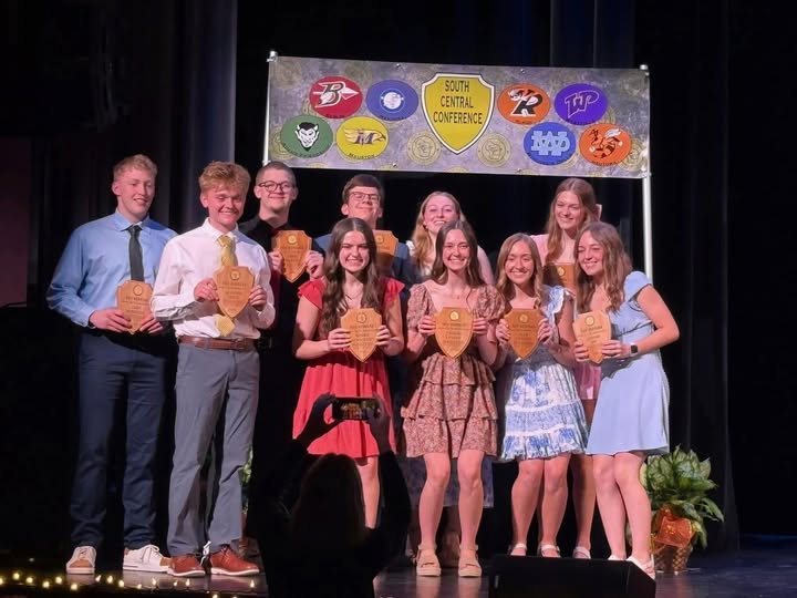 Ten students on a stage holding wooden plaques under a South Central Conference banner.