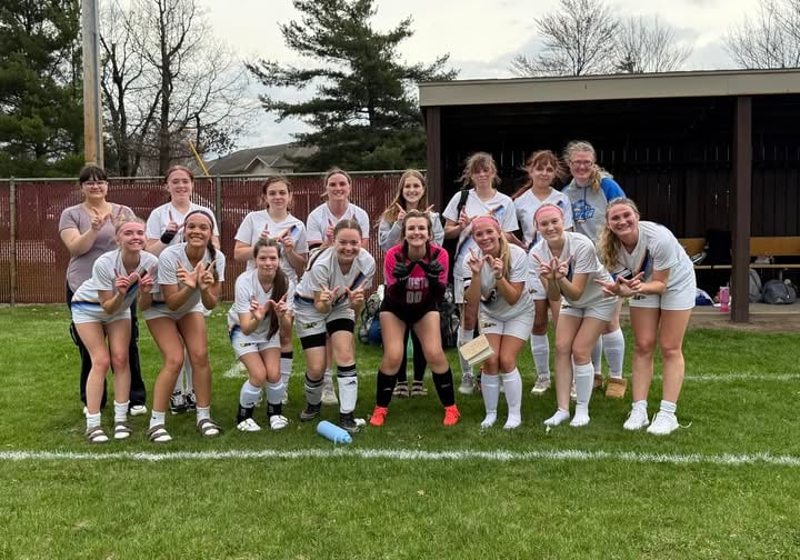 The girls' soccer team in white uniforms posing and making hand signs on a grass field.