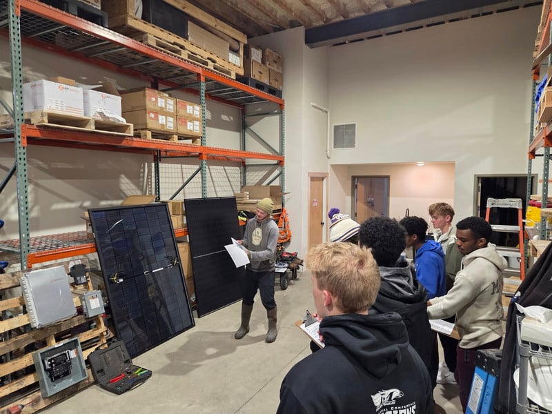 Students in a warehouse watching a demonstration of solar panels and electrical equipment.