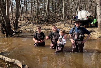 Four students wearing waterproof waders stand in a shallow river surrounded by trees.