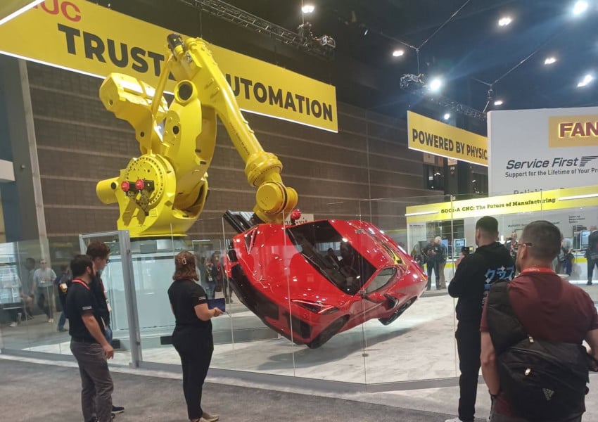 Students watch a large yellow FANUC industrial robot lifting a red sports car inside a display booth