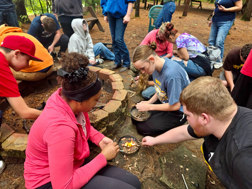 Students kneel outdoors around a fire pit learning how to start small fires with tinder pans.